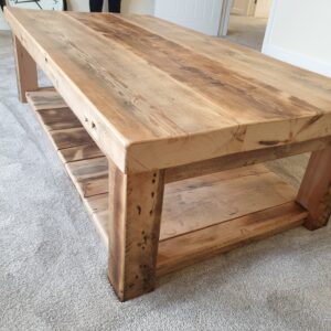 A close-up of a rustic wood coffee table, highlighting the rich, natural grain and unique imperfections of the reclaimed timber. The photo shows the table in a living room setting, with a floral arrangement on its surface, emphasizing its role as a functional and decorative centerpiece.
