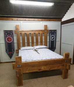 A unique, heavy-duty medieval bed frame crafted from 200-year-old reclaimed timber, featuring large, hand-forged steel bolts and brackets for a rustic, rugged appearance.
