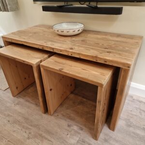 A rustic nest of tables made from reclaimed wood is pictured from a side angle, showcasing the sturdy, handcrafted joinery and the distinct patterns of the timber. The varying sizes of the tables are clearly visible, illustrating their clever design as a set.